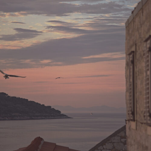 A pink and lavender Adriatic dusk seen from a stone window on the island of Vis, Croatia, with two gulls in flight and a distant fishing boat — limited edition fine art photograph by Tanja, My Empirical Life.