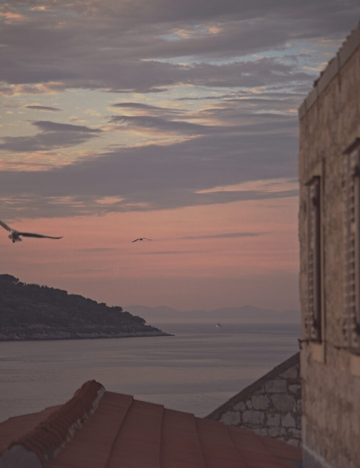 A pink and lavender Adriatic dusk seen from a stone window on the island of Vis, Croatia, with two gulls in flight and a distant fishing boat — limited edition fine art photograph by Tanja, My Empirical Life.