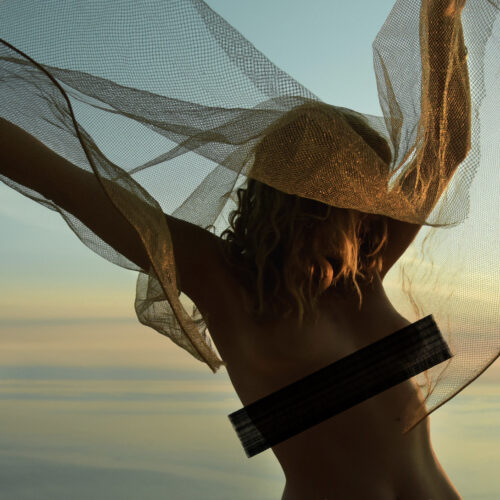 A blindfolded female figure stands at the edge of the sea with a translucent gold mesh veil stretched between her arms like wings, photographed during the pandemic — limited edition fine art photograph by Tanja, My Empirical Life. Listing image includes a display-only censor band; the print itself is unaltered.