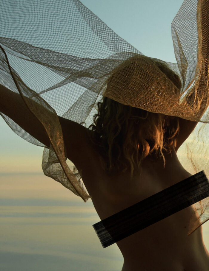 A blindfolded female figure stands at the edge of the sea with a translucent gold mesh veil stretched between her arms like wings, photographed during the pandemic — limited edition fine art photograph by Tanja, My Empirical Life. Listing image includes a display-only censor band; the print itself is unaltered.