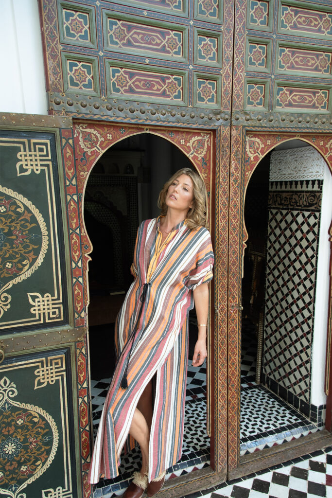A woman wearing a striped dress leans on an ornate moroccan door at Raid Kaiss by Anika in Marrakech